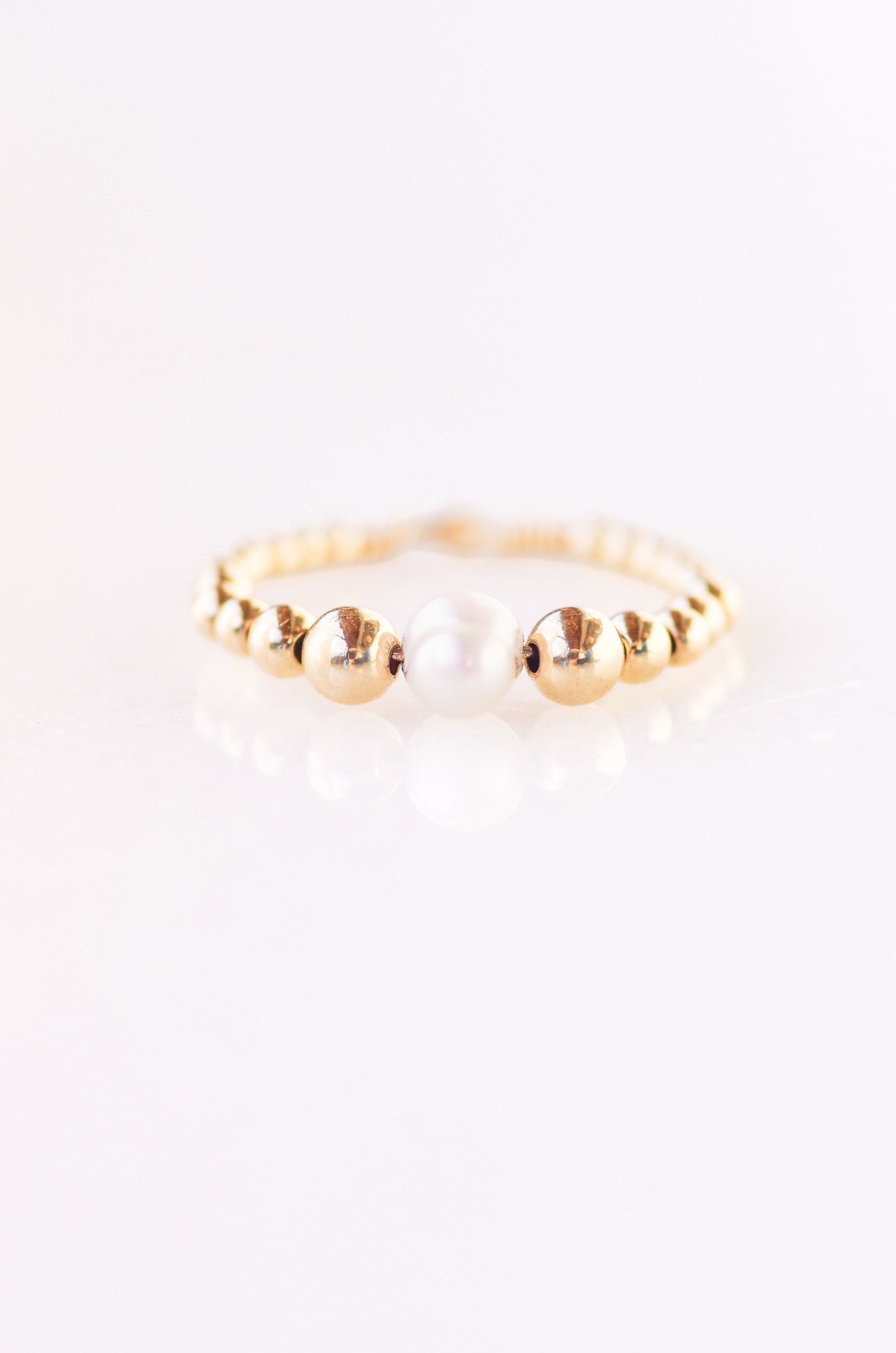Elegant beaded ring with a central 5mm irregular pearl, surrounded by 4mm, 3mm, 2.5mm, and 2mm gold beads, threaded on fine wire, displayed on a plain background almost white with violet hue