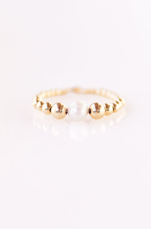Elegant beaded ring with a central 5mm irregular pearl, surrounded by 4mm, 3mm, 2.5mm, and 2mm gold beads, threaded on fine wire, displayed on a plain background almost white with violet hue