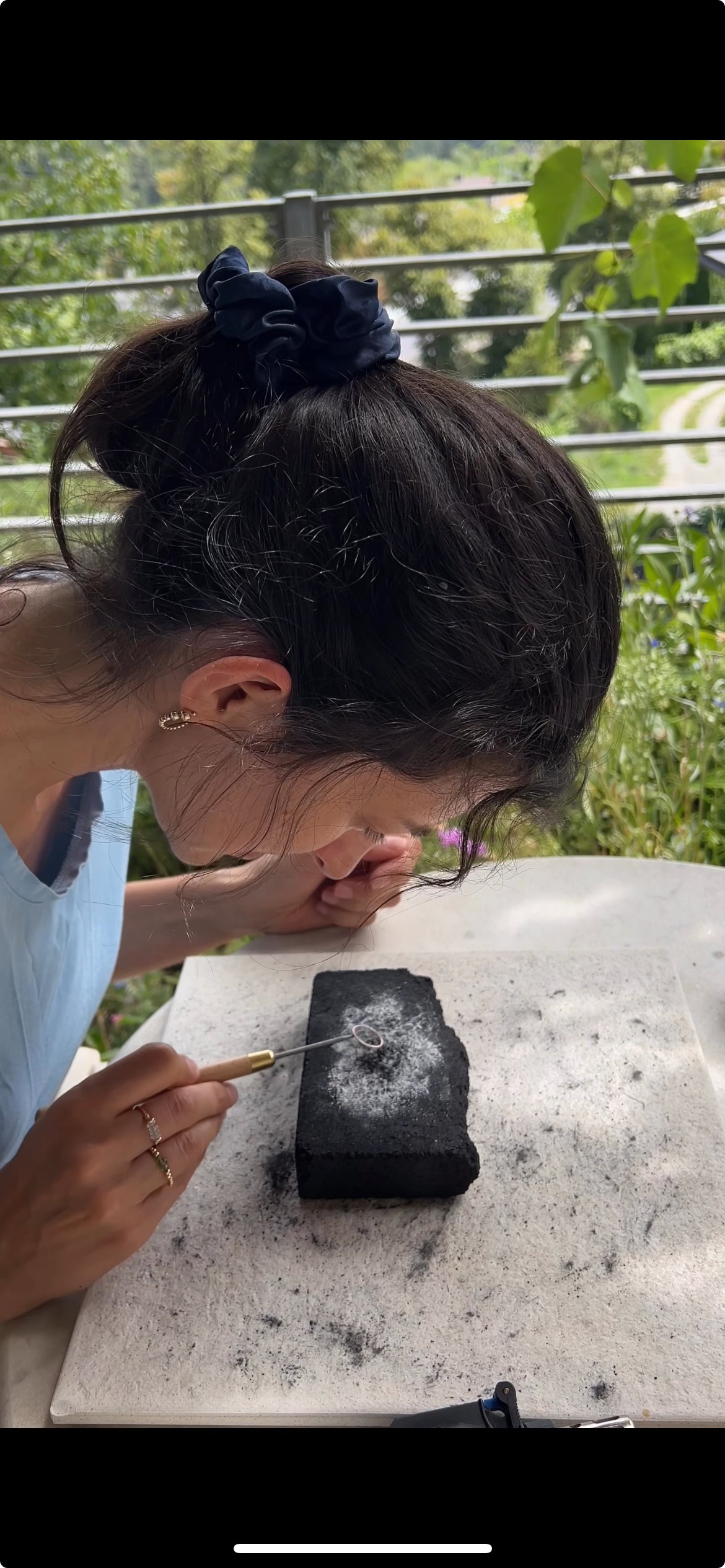 A woman with her hair in a bun leans over a small black rectangular object on a white surface, holding a fine brush in an outdoor setting with greenery and a metal railing in the background.