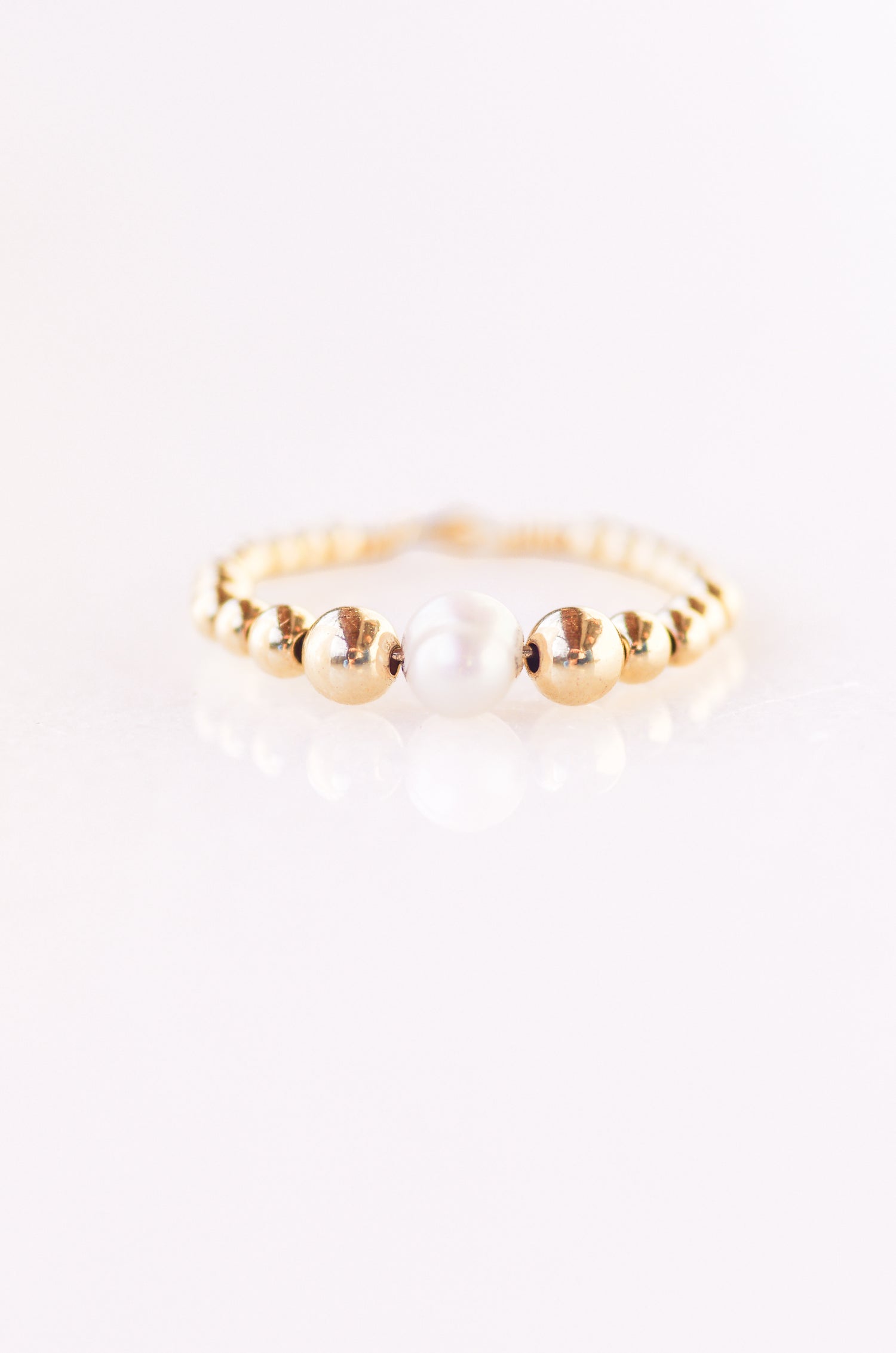 Elegant beaded ring with a central 5mm irregular pearl, surrounded by 4mm, 3mm, 2.5mm, and 2mm gold beads, threaded on fine wire, displayed on a plain background almost white with violet hue
