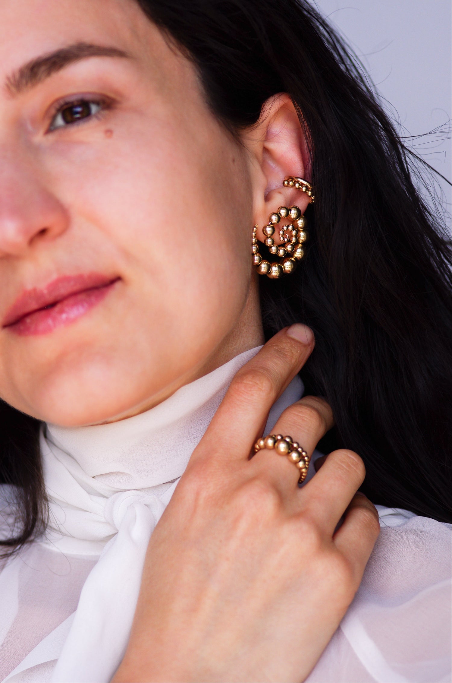 Woman wearing handcrafted gold spiral stud earrings with graduated beads, two modern ear cuffs stacked on the conch, and beaded gold rings, stacked. Spiral earrings are about 3cm wide and feature increasing bead sizes. Jewelry shown in a modern, elegant styling for pierced ears and stacked ring look. 